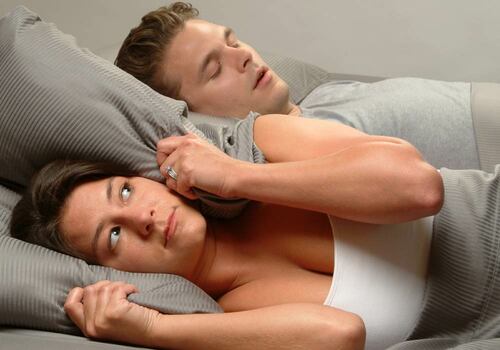 woman lying awake at night frustrated because husband is snoring; pillow around womans ears